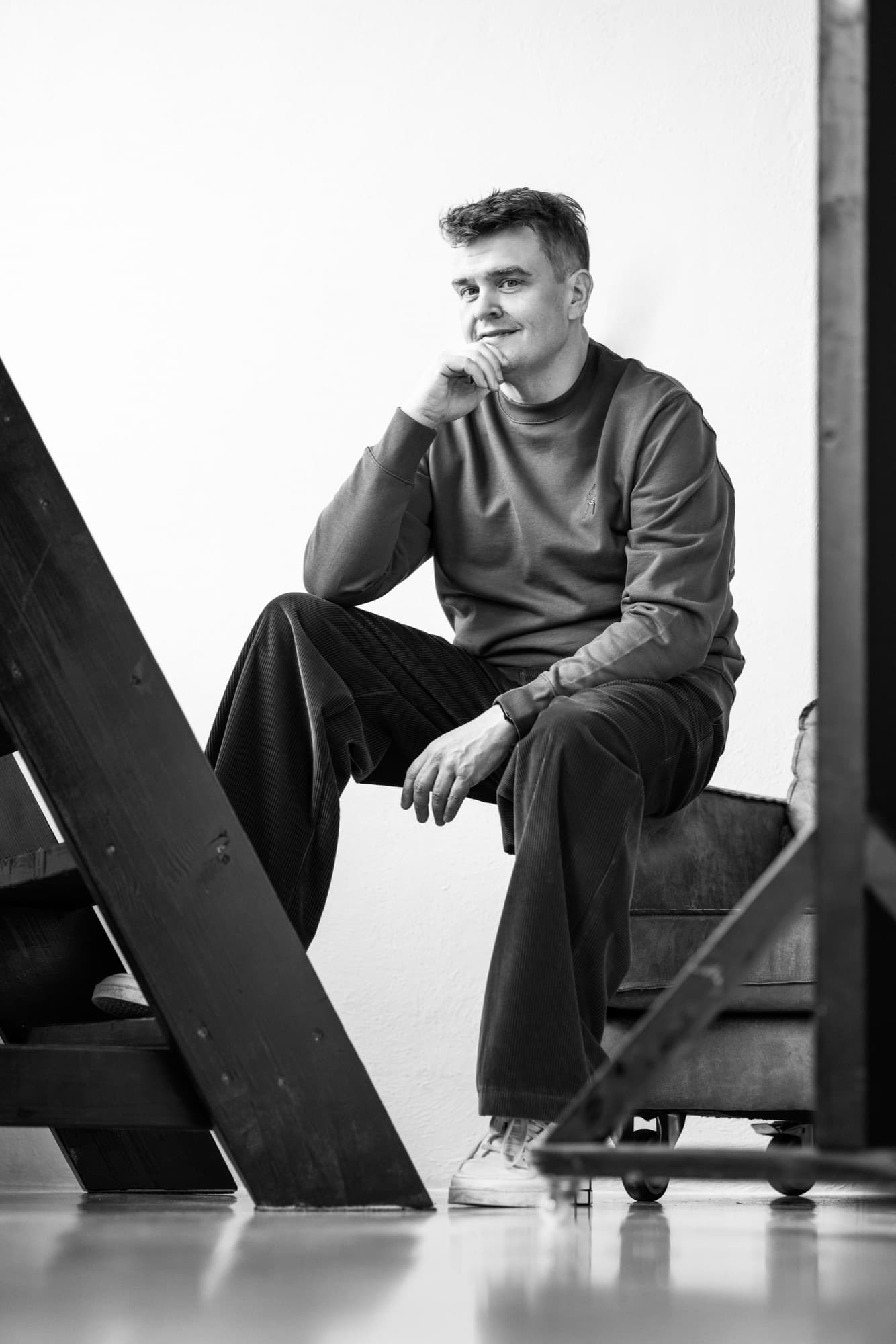A man sits on a wooden ladder-like structure in an industrial interior, chin resting on his hand, a small smile on his face.