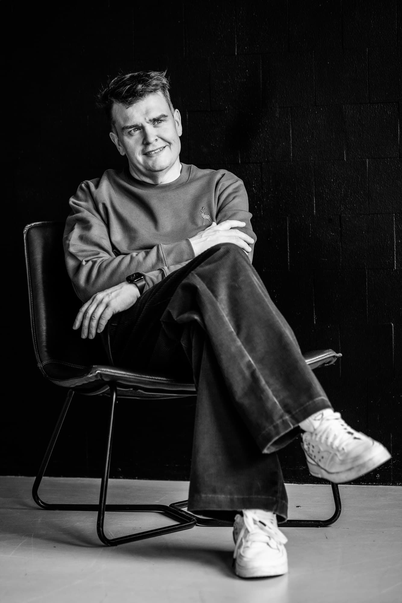 Mustavalkokuva miehestä, joka istuu tuolissa jalat ristissä ja kädet ristissä, yllään harmaa collegepaita ja tummat housut sekä valkoiset lenkkarit, hymyillen tummaa taustaa vasten.