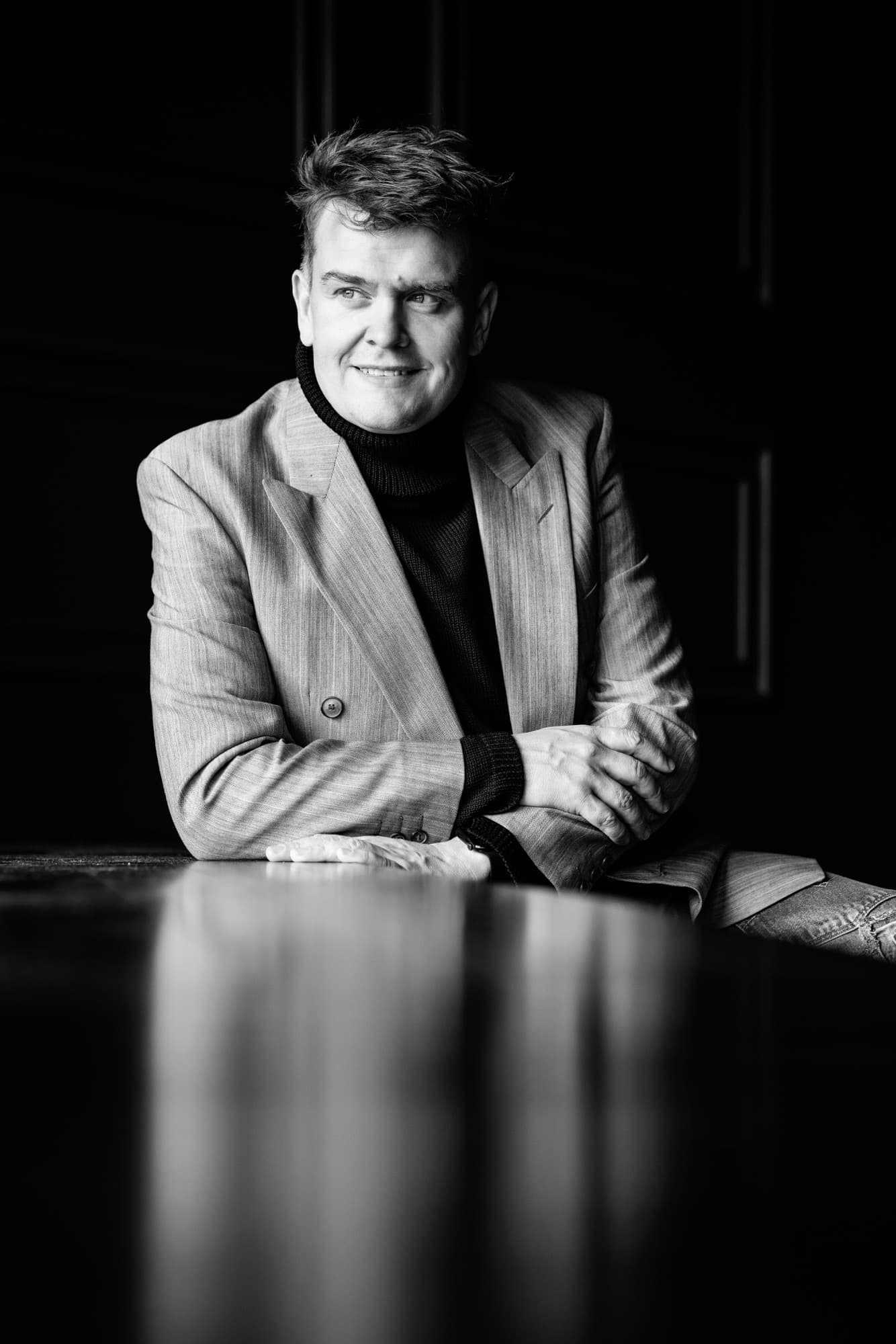 Black-and-white photo of a man leaning with his arms crossed on a dark glossy table, wearing a beige blazer and a black polo shirt, looking off to the side with a serious, thoughtful expression.
