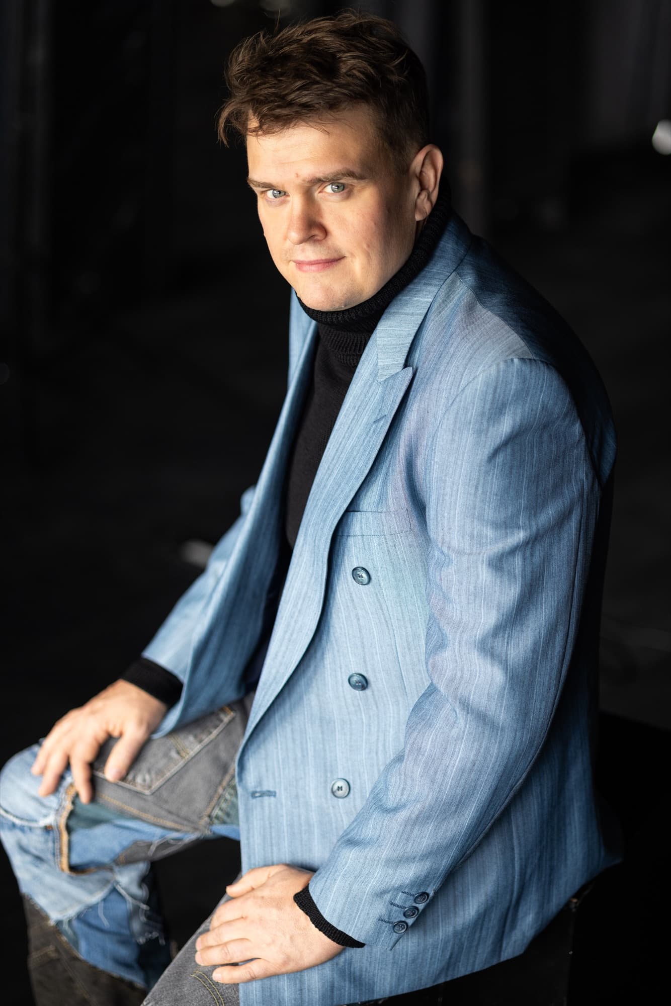 A man sits sideways on a stool, wearing a light blue blazer, a black polo shirt and ripped jeans, hand on his knee, looking back over his shoulder at the camera against a dark background.