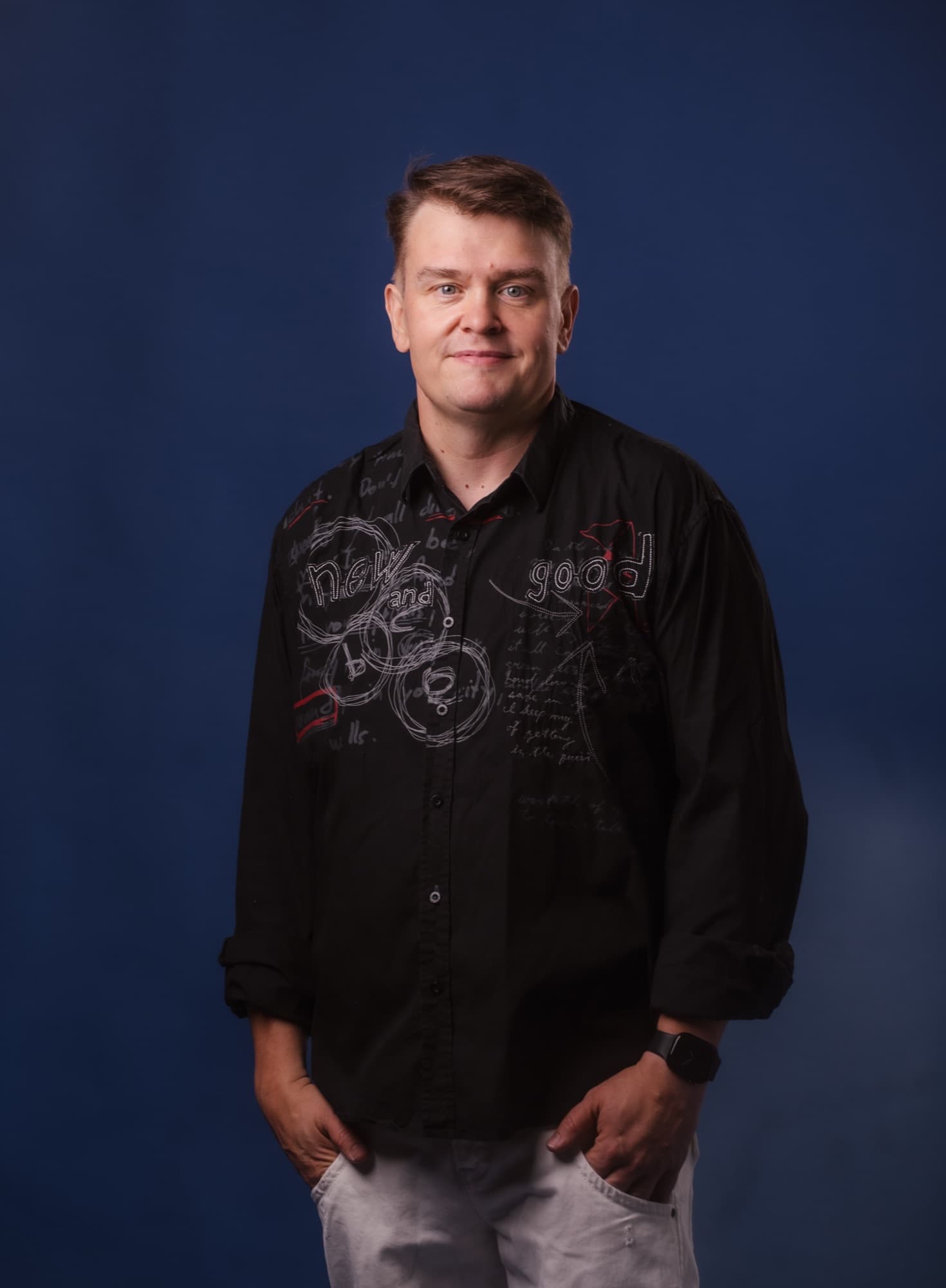 A man stands against a dark blue background wearing a black shirt with a text pattern and grey trousers, hands in his pockets, looking straight at the camera with a slight smile.