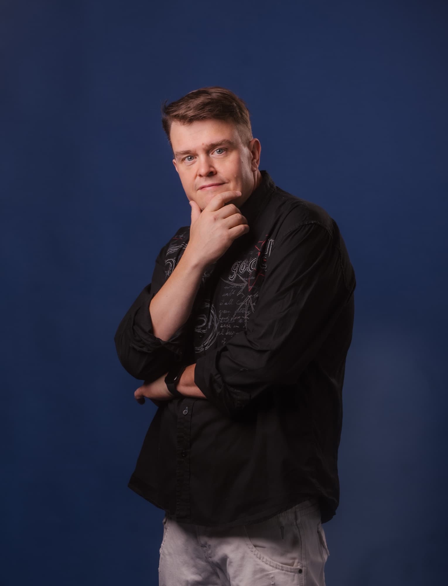A studio photo of a man against a dark blue background, standing sideways and touching his chin in a thoughtful pose.